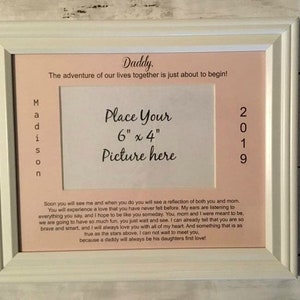May include: A white picture frame with a pink mat and a 6x4 inch opening for a photo. The frame has the text "Daddy. The adventure of our lives together is just about to begin!" and "Place Your 6" x 4" Picture here" printed on it. The frame also has the text "Madison" and "2019" printed vertically on the left and right sides of the mat.