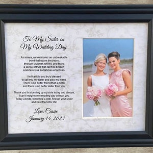 May include: A framed wedding day poem for a sister, with a photo of two women in wedding attire. The poem reads "To My Sister on My Wedding Day" and includes the text "As sisters, we've shared an unbreakable bond that spans the years, through laughter, smiles, and tears; a sense of trust that can't be broken, a sincere love sometimes unspoken." The poem concludes with "Love, Cassie January 14, 2023".