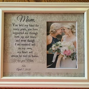 May include: A framed photo with a white frame and a white mat. The photo shows two women, one in a white dress and the other in a blue dress, smiling at each other. The photo is accompanied by a poem that reads "Mom, You held my hand for many years, you have supported me through both joy and tears, and even though I am married and on my own, my heart will always be tied to home. I love you Mom. Abi April 7, 2020."