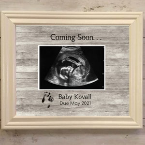 May include: A framed ultrasound image with the text "Coming Soon..." and "Baby Kovall Due May 2021". The frame is off-white, and the background has a wood-grain pattern.