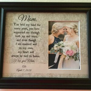 May include: A framed photo with a message to a mother from her daughter. The message reads "Mom, You held my hand for many years, you have supported me through both joy and tears, and even though I am married and on my own, my heart will always be tied to home. I love you Mom. Abi April 7, 2020". The photo shows two women, likely a mother and daughter, smiling at each other. The daughter is wearing a white dress and the mother is wearing a dark blue dress. They are holding a bouquet of flowers.