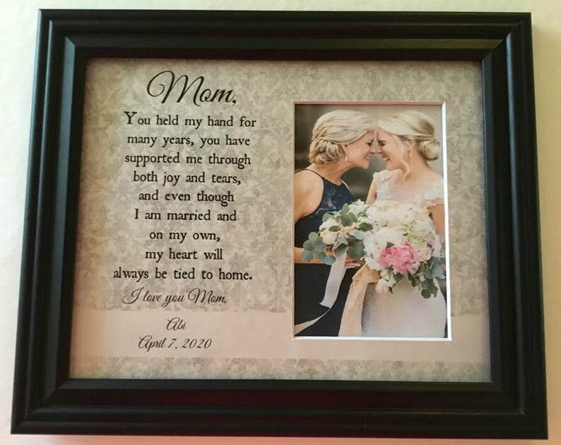 May include: A framed photo with a message to a mother from her daughter. The message reads "Mom, You held my hand for many years, you have supported me through both joy and tears, and even though I am married and on my own, my heart will always be tied to home. I love you Mom. Abi April 7, 2020". The photo shows two women, likely a mother and daughter, smiling at each other. The daughter is wearing a white dress and the mother is wearing a dark blue dress. They are holding a bouquet of flowers.