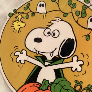 May include: A circular cardboard cutout featuring Snoopy dressed as a vampire with a black cape and fangs, standing behind an orange jack-o'-lantern. The background is yellow with green vines, a small yellow bird hanging from a branch, and a white ghost.