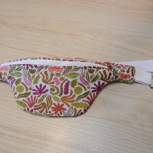 May include: A white fanny pack with a zipper closure and an adjustable strap. The fabric has a colorful floral pattern with pink, orange, green, and brown accents.