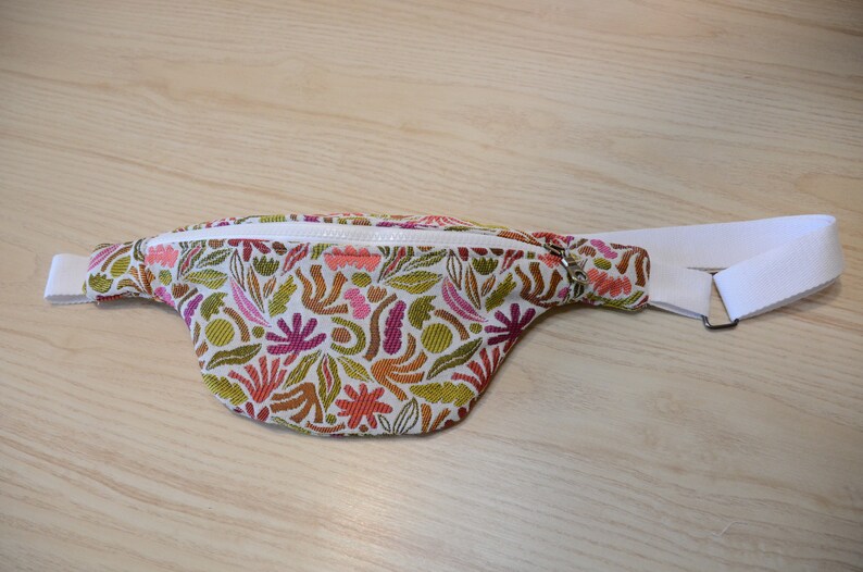 May include: A white fanny pack with a zipper closure and an adjustable strap. The fabric has a colorful floral pattern with pink, orange, green, and brown accents.