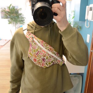 May include: A white fanny pack with a colorful floral print. The fanny pack is worn across the body and has a zipper closure.