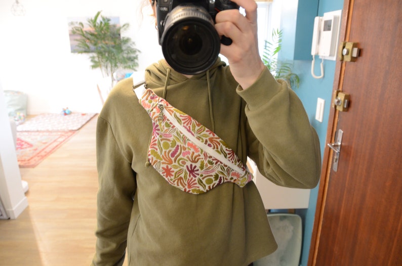 May include: A white fanny pack with a colorful floral print. The fanny pack is worn across the body and has a zipper closure.