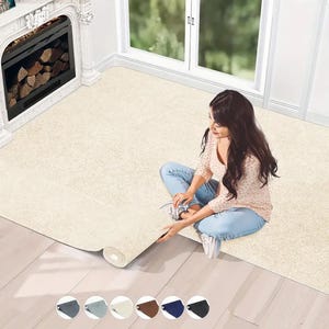 May include: A cream-colored rectangular rug is being unrolled on a hardwood floor. The rug is a neutral color and appears to be made of a soft material. A woman is sitting on the floor, unrolling the rug. A fireplace and window are in the background.