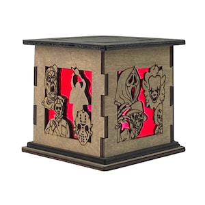 Puede incluir: Lámpara cúbica de madera con diseños de personajes de películas de terror cortados con láser. La lámpara tiene una parte superior e inferior oscuras, con luz roja que ilumina los personajes desde el interior. Los personajes son de películas de terror icónicas.