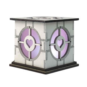 Puede incluir: Una caja de madera blanca con un brillo púrpura claro y dos recortes en forma de corazón. La caja tiene una base marrón oscuro y una tapa marrón oscuro.