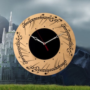 Puede incluir: Un reloj redondo de madera con una esfera negra y agujas blancas. La esfera del reloj está rodeada por un anillo de texto negro en una fuente de escritura. El texto dice "Un anillo para gobernarlos a todos, Un anillo para encontrarlos, Un anillo para atraerlos a todos y en las tinieblas atarlos." El reloj está situado sobre un fondo de un paisaje de fantasía.