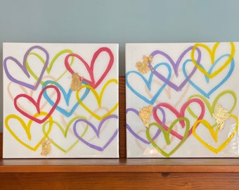 May include: Two square canvases with a white background and a colorful design of overlapping hearts in shades of yellow, green, pink, blue, and purple. The hearts are outlined in black and have gold accents.
