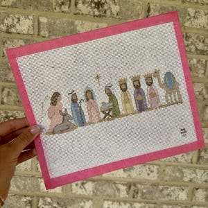 Könnte beinhalten: Eine rechteckige Stickleinwand mit einer Krippenszene, die einen Engel, einen Esel, Josef, Maria, das Jesuskind in der Krippe, drei Könige und ein Kamel zeigt. Das Motiv ist von einem leuchtend rosa Stoffrand auf weißem Gitter umgeben.