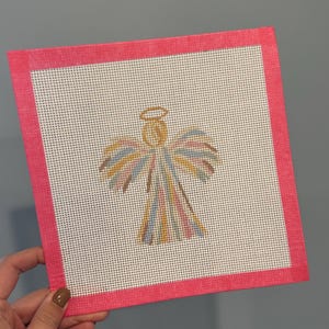 May include: A square needlepoint canvas featuring a central angel design stitched in multicolored pastel threads with a gold halo. The canvas is framed by a bright pink fabric border on all four sides, creating a distinct contrast.