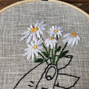 May include: Embroidered daisy bouquet in a wooden hoop. The white daisies with yellow centers are held by a hand drawn in black thread. The fabric is a light tan color.