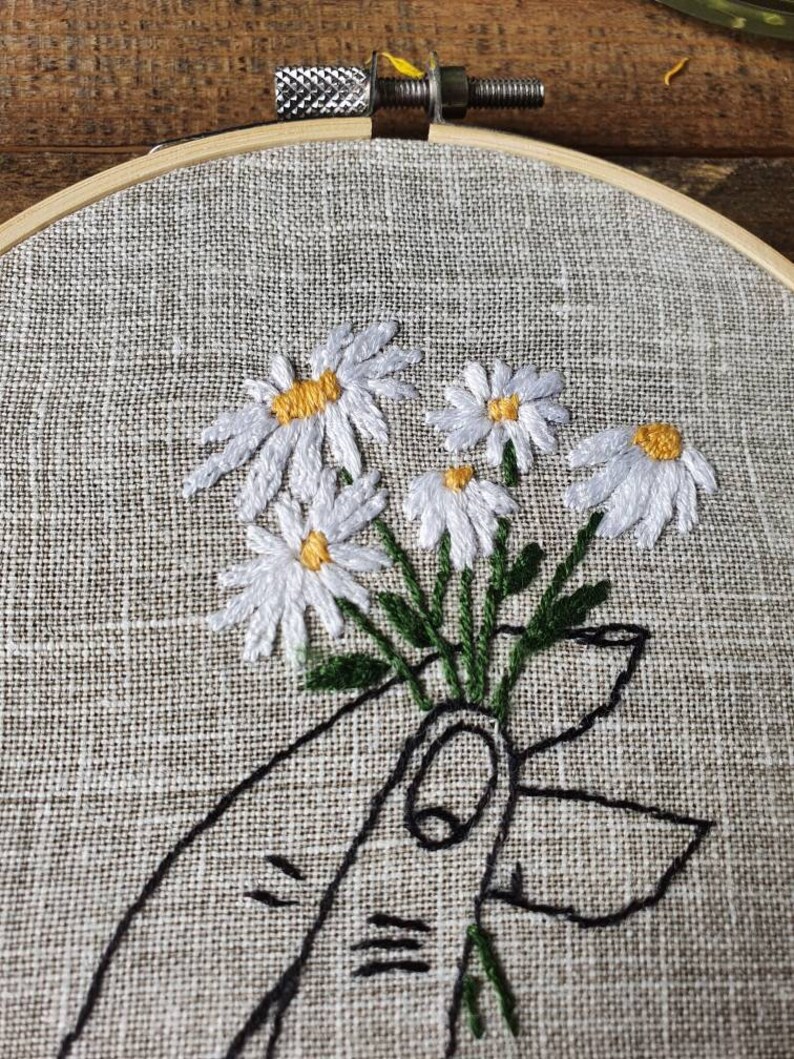 May include: Embroidered daisy bouquet in a wooden hoop. The white daisies with yellow centers are held by a hand drawn in black thread. The fabric is a light tan color.