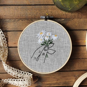 May include: A hand-stitched embroidery hoop featuring a bouquet of white daisies with yellow centers. The flowers are held by a hand, all embroidered with black thread on a gray linen fabric. The wooden hoop is visible, and the background is a wooden surface.