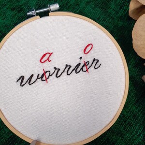May include: A white fabric stretched within a wooden embroidery hoop. The text "a worrier" is stitched in black thread, with the letters "a" and "o" in red, crossed out with red thread. The background is a textured green fabric.