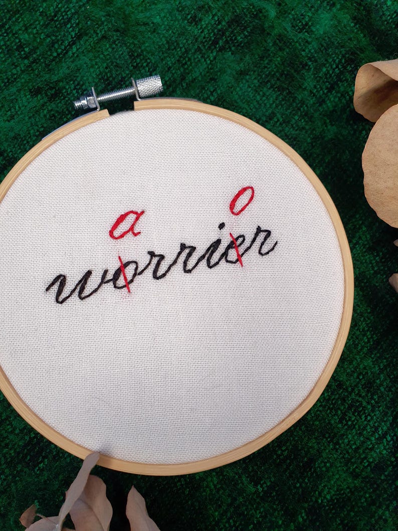 May include: A white fabric stretched within a wooden embroidery hoop. The text "a worrier" is stitched in black thread, with the letters "a" and "o" in red, crossed out with red thread. The background is a textured green fabric.