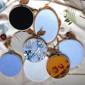 May include: A collection of fabric-covered embroidery hoops in various sizes and designs. The hoops feature patterns like polka dots, ballet dancers, and floral prints. Each hoop is adorned with a small tag, and the color palette includes blue, yellow, and neutral tones.
