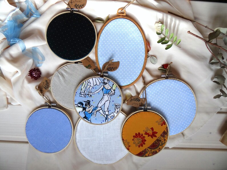 May include: A collection of fabric-covered embroidery hoops in various sizes and designs. The hoops feature patterns like polka dots, ballet dancers, and floral prints. Each hoop is adorned with a small tag, and the color palette includes blue, yellow, and neutral tones.
