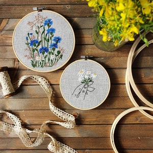 May include: Two embroidered floral hoops with blue and white flowers, and a hand holding daisies. A spool of thread and lace ribbon are on a wooden surface. Yellow flowers in a glass vase are in the background.