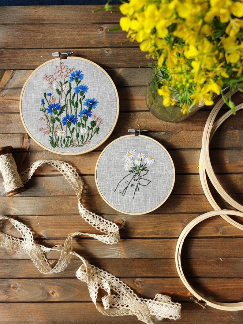May include: Two embroidered floral hoops with blue and white flowers, and a hand holding daisies. A spool of thread and lace ribbon are on a wooden surface. Yellow flowers in a glass vase are in the background.