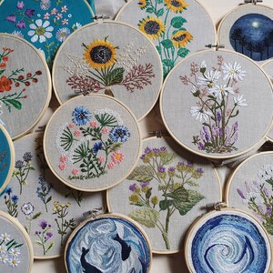 May include: A collection of embroidered floral designs displayed in wooden hoops. The embroidery features various flowers, including sunflowers, daisies, and wildflowers, in vibrant colors. The hoops are arranged on a neutral linen background, showcasing the handmade craft.