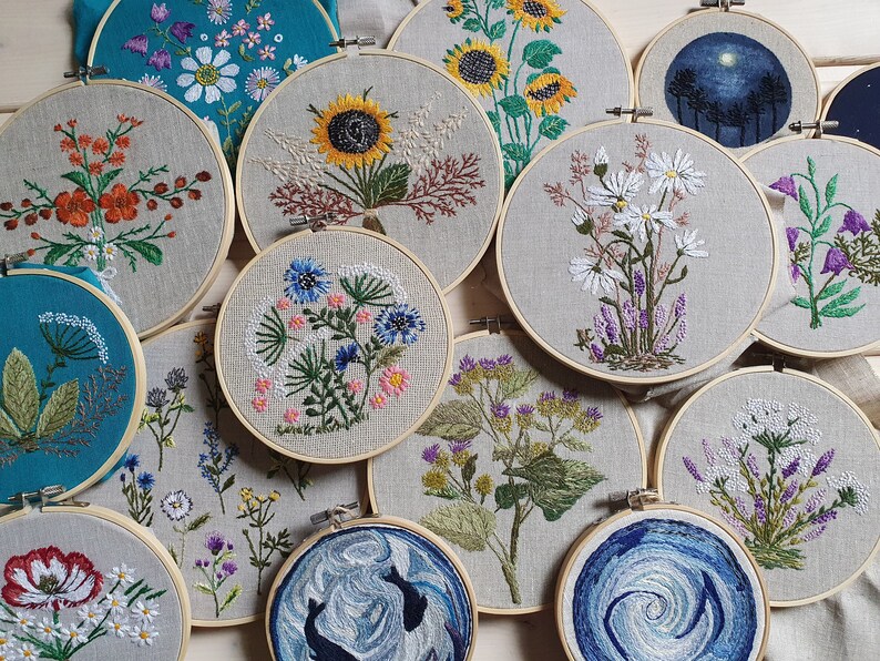 May include: A collection of embroidered floral designs displayed in wooden hoops. The embroidery features various flowers, including sunflowers, daisies, and wildflowers, in vibrant colors. The hoops are arranged on a neutral linen background, showcasing the handmade craft.