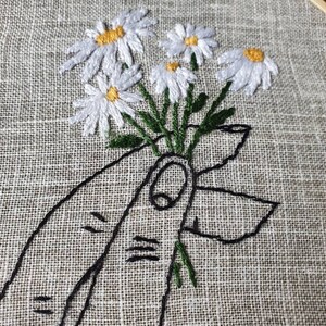 May include: Embroidered artwork featuring a bouquet of white daisies with yellow centers and green stems, held by a black outline of a hand. The embroidery is set in a wooden hoop against a textured, neutral-toned fabric.
