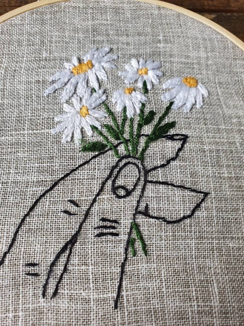 May include: Embroidered artwork featuring a bouquet of white daisies with yellow centers and green stems, held by a black outline of a hand. The embroidery is set in a wooden hoop against a textured, neutral-toned fabric.