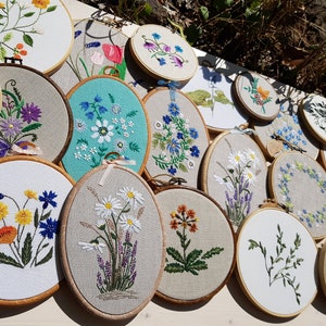 May include: A collection of embroidered floral designs displayed on wooden hoops. The embroidery features various flowers in vibrant colors, including daisies, lavender, and sunflowers, on a neutral fabric background. The hoops are arranged on a white surface.
