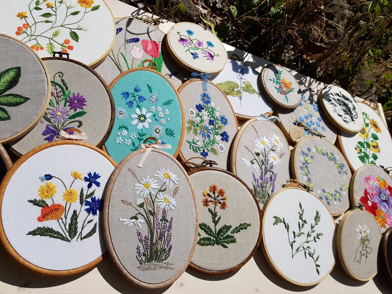 May include: A collection of embroidered floral designs displayed on wooden hoops. The embroidery features various flowers in vibrant colors, including daisies, lavender, and sunflowers, on a neutral fabric background. The hoops are arranged on a white surface.