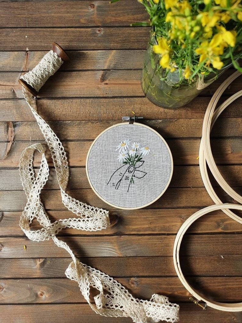 May include: An embroidery hoop with a hand holding daisies, a spool of lace ribbon, and additional embroidery hoops are arranged on a wooden surface. A glass jar with yellow flowers is in the background. The embroidery features white flowers with green stems.