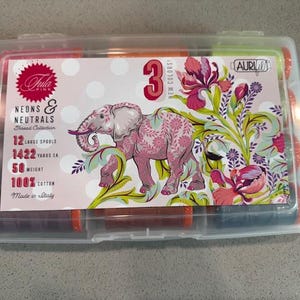 May include: A clear plastic container holding 12 spools of thread from the Tula Pink Neons & Neutrals collection. The label features a pink elephant illustration and floral accents. Each spool contains 1422 yards of 100% cotton thread.
