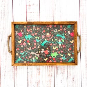 May include: Rectangular wooden serving tray with a dark background and a colorful pattern of whimsical figures, animals, and floral elements. The tray has woven handles on each side and a wooden frame.