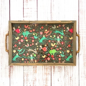 May include: Rectangular wooden serving tray with a decorative, colorful pattern. The pattern features whimsical illustrations of animals, figures, and floral elements in shades of green, red, and pink on a dark background. The tray has woven handles.