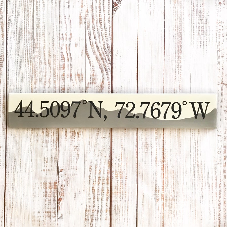 May include: A wooden sign with a light brown background and a dark gray mountain silhouette. The sign displays the coordinates "44.5097&deg;N, 72.7679&deg;W" in black text.