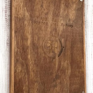 Puede incluir: Una tabla rectangular de madera con un acabado marr&oacute;n oscuro. La tabla presenta texto grabado y un emblema circular. El texto dice "1838 7503 Topographical Survey". El emblema incluye las letras "OR" dentro de un dise&ntilde;o de br&uacute;jula.