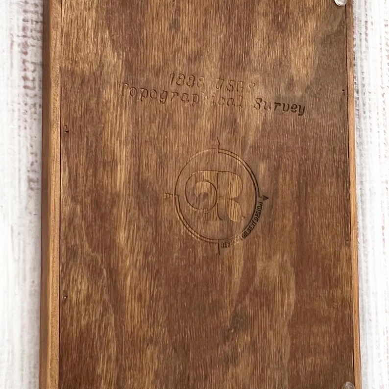 Puede incluir: Una tabla rectangular de madera con un acabado marr&oacute;n oscuro. La tabla presenta texto grabado y un emblema circular. El texto dice "1838 7503 Topographical Survey". El emblema incluye las letras "OR" dentro de un dise&ntilde;o de br&uacute;jula.