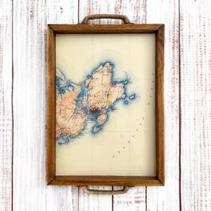 May include: Rectangular wooden serving tray with a vintage map design. The map features shades of blue and orange, with the words "Atlantic Ocean" along the right side. The tray has rope handles and a light wood frame.