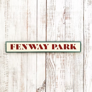 May include: A white wooden sign with green trim and the words "Fenway Park" in red lettering.