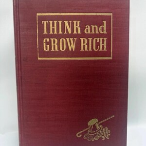 Może przedstawiać: Książka w twardej oprawie o głębokim czerwonym, teksturowanym kolorze. Tytuł THINK and GROW RICH jest wydrukowany złotymi literami w prostokątnej ramce. Poniżej tytułu mała złota grafika przedstawia cylinder, laskę oraz stos monet.