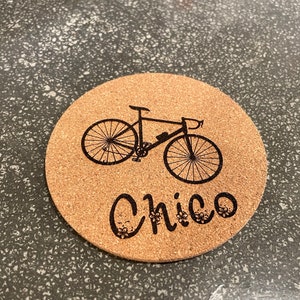 Puede incluir: Un posavasos de corcho con una silueta negra de una bicicleta y la palabra "Chico" impresa en él.