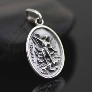 May include: Silver pendant featuring a detailed depiction of Saint Michael the Archangel slaying a dragon. The pendant is oval shaped and has the words "St. Michael" inscribed on the side.