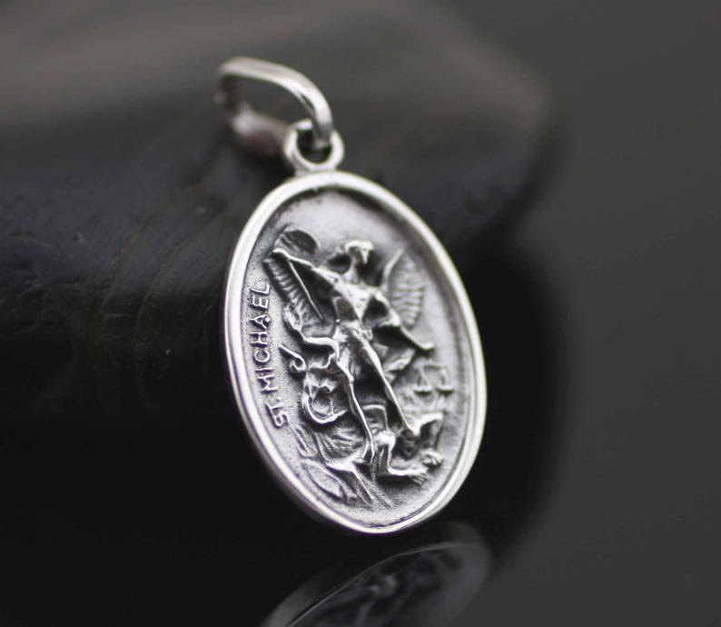 May include: Silver pendant featuring a detailed depiction of Saint Michael the Archangel slaying a dragon. The pendant is oval shaped and has the words "St. Michael" inscribed on the side.
