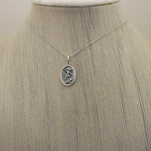 May include: A silver chain necklace with a silver oval pendant featuring a detailed depiction of an angel with wings outstretched.