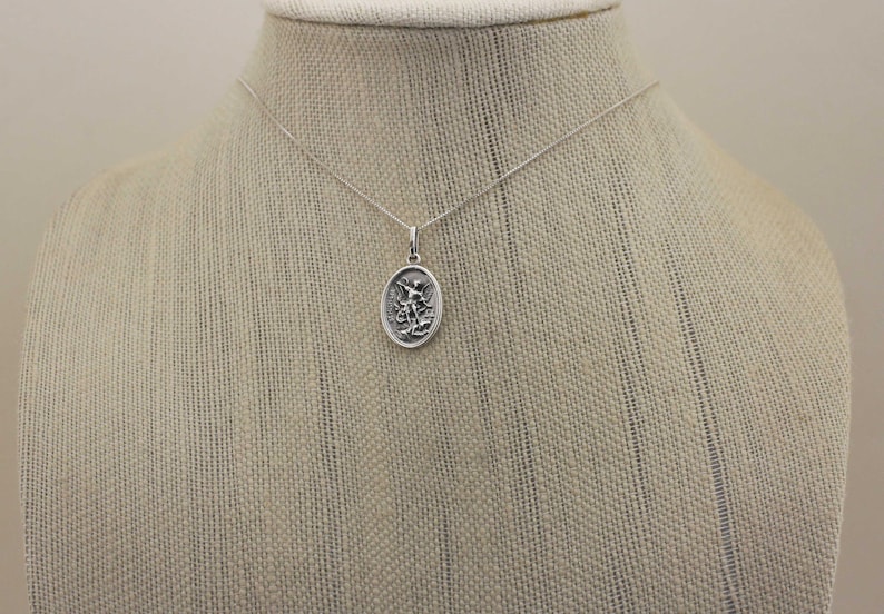 May include: A silver chain necklace with a silver oval pendant featuring a detailed depiction of an angel with wings outstretched.