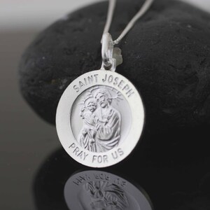 May include: Silver pendant necklace with a round charm featuring a depiction of Saint Joseph holding baby Jesus. The charm is inscribed with "Saint Joseph Pray For Us".
