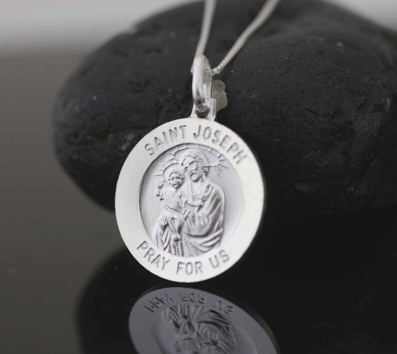 May include: Silver pendant necklace with a round charm featuring a depiction of Saint Joseph holding baby Jesus. The charm is inscribed with "Saint Joseph Pray For Us".